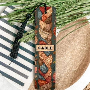 May include: A bookmark featuring a colorful cable knit pattern in shades of blue, orange, brown, and cream. The bookmark has a dark green background and the word "CABLE" printed in white on a brown rectangle.