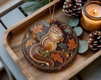 Pode incluir: Um ornamento circular, plano e redondo, com uma ilustração detalhada de um esquilo-listrado segurando uma bolota, rodeado por folhas de carvalho em tons de outono e pequenas bolotas sobre um fundo escuro, suspenso por um fio dourado fino.