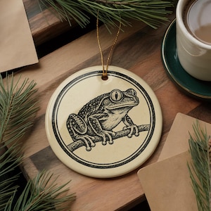 May include: A round, cream-colored ceramic ornament featuring a black ink-style illustration of a frog perched on a branch. The design is enclosed within a circular border, and a thin gold string is attached to the top for hanging purposes.