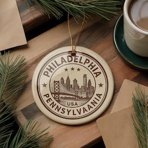 May include: Cream-colored round ornament with a brown Philadelphia, Pennsylvania, USA design. The design features a bridge, city skyline, stars, and text. The ornament has a gold string for hanging.