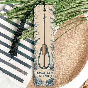 May include: A rectangular bookmark with a cream background featuring a blue illustration of a Hawaiian sling spear fishing tool. The design includes underwater coral and seaweed motifs, a black tassel at the top, and the text HAWAIIAN SLING.