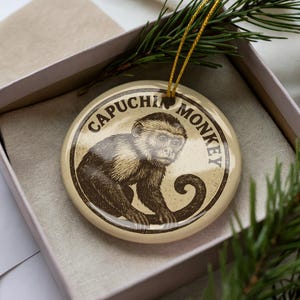 May include: A round, cream-colored ornament featuring a detailed illustration of a capuchin monkey. The text "CAPUCHIN MONKEY" is printed around the edge in a dark brown color. The ornament is in a gift box with a gold string for hanging.