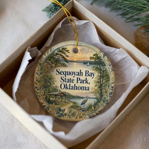 May include: A round ceramic ornament featuring a scenic illustration of Sequoyah Bay State Park, Oklahoma. The design includes a lake, trees, and a heron, with the park's name in blue text. The ornament is presented in a beige gift box with tissue paper.