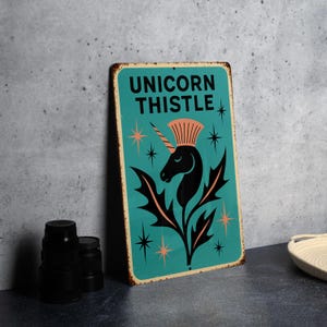 May include: A teal metal sign with the words "UNICORN THISTLE" in white. The sign features a black unicorn head with a crown and thistle leaves. Orange starbursts and a distressed border complete the design.