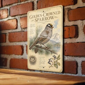 May include: A vintage-style metal sign featuring a Golden-Crowned Sparrow illustration. The sign includes the bird's scientific name, field notes, and a depiction of the bird perched on a pine branch with a pine cone and berries. The sign is mounted on a brick wall.