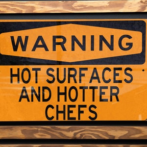 May include: A rectangular orange warning sign with black text that reads "WARNING HOT SURFACES AND HOTTER CHEFS." The sign is mounted on a wooden slatted wall.