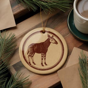 May include: A round, cream-colored ceramic ornament featuring a brown illustration of an okapi within a circular frame. The okapi is depicted in profile, with striped legs and a long neck. A gold string is attached for hanging.