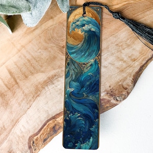 May include: A wooden bookmark featuring a detailed illustration of a wave with shades of blue and gold. The bookmark has a black tassel attached to the top. The design is reminiscent of Japanese art.