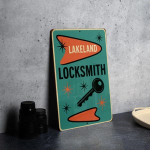 May include: A vintage-style metal sign with a teal background and orange accents. The sign reads "LAKELAND LOCKSMITH" in black text, with a key graphic. Decorative starbursts surround the text and key, giving it a retro look.