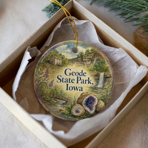 May include: A round ceramic ornament featuring a scenic illustration of Geode State Park, Iowa, with a waterfall, bridge, and amethyst geodes. The ornament is in a gift box with tissue paper and a gold string.