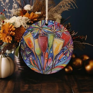 May include: Round ceramic ornament with a colorful design of five champagne flutes against a background of fireworks and confetti.