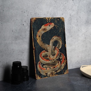 May include: A rectangular metal sign featuring a coiled snake illustration. The snake has a red head, tan body with black markings, and red and white accents. The background is dark blue with gold decorative elements.