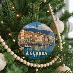 May include: A round ceramic ornament hanging on a Christmas tree, featuring a colorful coastal scene of A Guarda, Spain, with buildings, boats, and water. The text A GUARDA SPAIN is printed in white at the bottom of the circular design.