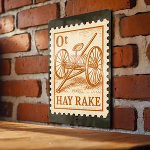 May include: A rectangular metal sign with a vintage design resembling a postage stamp. The sign features an illustration of a hay rake and the text "HAY RAKE" in a serif font. The background is a brick wall.