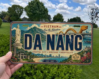 May include: A rectangular metal license plate featuring a colorful illustration of Da Nang, Vietnam. The design includes a statue, a golden dragon bridge, and coastal scenery with the text "VIETNAM Live the Adventure", "DA NANG", "COASTAL CITY HERITAGE HOSPITALITY", "BÀ NÀ HILLS", and "EST. 1997".