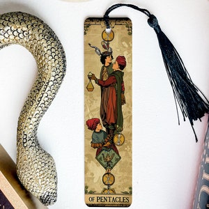May include: A bookmark featuring a vintage illustration of the Ten of Pentacles tarot card. The card depicts three figures, two men and a woman, with a golden coin and a bird in the center. The bookmark is made of wood and has a black tassel.