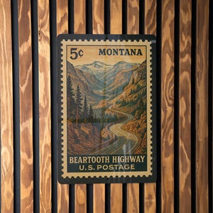 May include: A vintage-style metal sign resembling a postage stamp, featuring a scenic mountain landscape with a winding road. The sign reads "5¢ MONTANA" and "BEARTOOTH HIGHWAY U.S. POSTAGE."