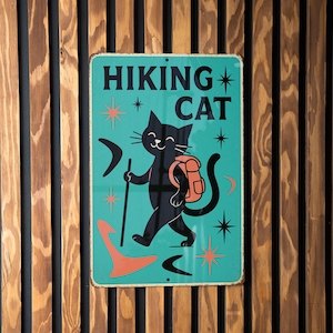 May include: A teal metal sign with the words "HIKING CAT" and a cartoon black cat wearing a backpack and holding a walking stick. The sign has a retro design with star and arrow accents, mounted on a wooden slat wall.