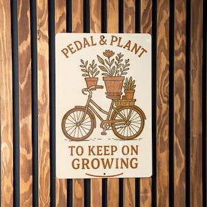 May include: A white metal sign with a brown bicycle carrying potted plants. The sign reads "Pedal & Plant to Keep on Growing."