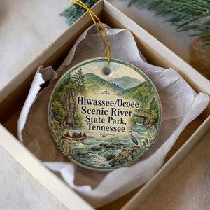 May include: A round ceramic ornament featuring a scenic river landscape with the text "Hiwassee/Ocoee Scenic River State Park, Tennessee." The ornament is in a gift box with tissue paper, and has a gold string for hanging.