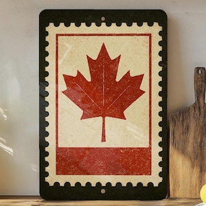 May include: A rectangular metal sign featuring a distressed design of a Canadian postage stamp. The stamp has a red maple leaf on a cream background, framed by a red border and a black scalloped edge. A hole is at the top for hanging.