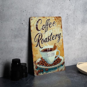 May include: A vintage-style metal sign with the text "Coffee Roastery" in brown lettering on a yellow background. A steaming cup of coffee with cinnamon sticks is on a blue and white plate with coffee beans. The text "Fresh Brew" is below the cup.
