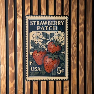 May include: A vintage-style metal sign featuring a strawberry patch design. The sign displays three ripe strawberries, white flowers, and the text "STRAWBERRY PATCH" and "USA 5c". The sign is mounted on a wooden slatted wall.