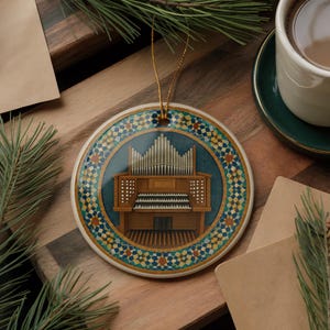 May include: Round Christmas ornament featuring a detailed illustration of a pipe organ. The ornament has a blue and gold geometric border and a gold hanging string. The background is a wooden surface with a cup of coffee and pine needles.