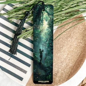 May include: A black bookmark with a green and gold illustration of a woman with a bow and arrow standing in a forest. The text "Magical Archer" is at the bottom of the bookmark.