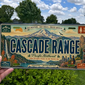 May include: A rectangular metal license plate featuring a vintage-style illustration of the Cascade Range. The design includes snow-capped mountains, a waterfall, a totem pole, and the text Cascade Range, Pacific Northwest, Oregon, Washington.