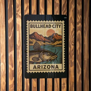 May include: A rectangular metal sign featuring a vintage-style illustration of a fish in a lake with mountains and a setting sun. The sign reads "BULLHEAD CITY ARIZONA" in a postage stamp design. The sign is mounted on a wooden wall.
