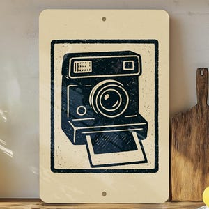 May include: A rectangular sign featuring a black and white illustration of a vintage instant camera. The camera is centered on a cream-colored background, with a black border. The sign has two small holes at the top and bottom.