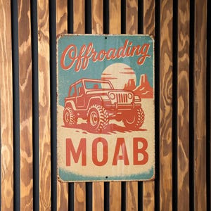 May include: Vintage-style metal sign with the words "Offroading MOAB" and a red Jeep illustration against a blue and tan background. The sign is mounted on a wooden slatted wall.
