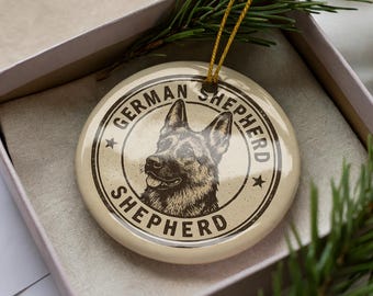 May include: A round, cream-colored ornament featuring a detailed illustration of a German Shepherd dog. The text "GERMAN SHEPHERD" is printed around the dog's image in a circular design. The ornament is in a gift box with a gold string.