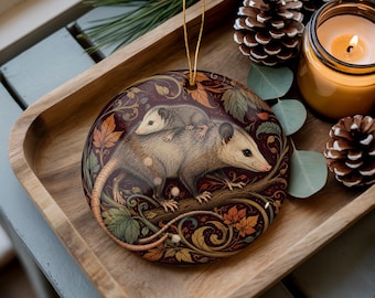 Pode incluir: Um ornamento de cerâmica redondo e plano com uma ilustração detalhada de um gambá adulto carregando um gambá menor nas costas. O design inclui um fundo bordô escuro com vinhas intrincadas e folhas de outono coloridas.