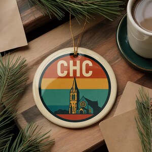 May include: A round ceramic ornament with a retro design featuring the text "CHC" in white, a church silhouette, and horizontal stripes in red, yellow, and teal. The ornament has a gold string for hanging.