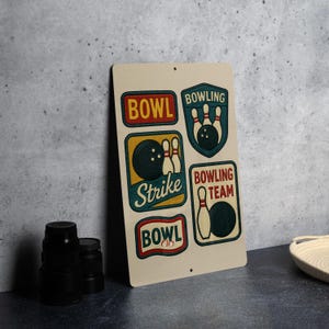 May include: A rectangular metal sign with a beige background features bowling-themed graphics. The sign includes the words "BOWL," "STRIKE," and "BOWLING TEAM" in various fonts and colors, along with bowling pin and ball illustrations. The sign has a vintage, retro aesthetic.