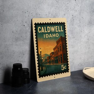A framed vintage postage stamp from Caldwell, Idaho, featuring an illustration of a street scene with buildings and a lamppost.