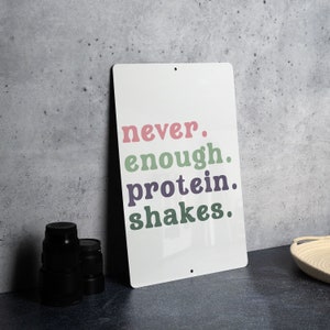 May include: White metal sign with the text "never. enough. protein. shakes." in colorful lettering. The sign is leaning against a gray concrete wall.