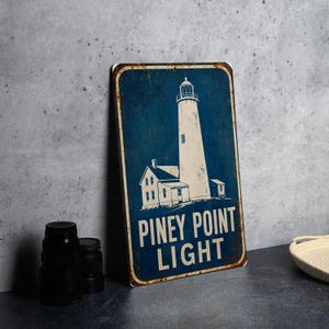 May include: A rectangular metal sign with a distressed blue finish featuring a white illustration of a lighthouse and the text PINEY POINT LIGHT. The sign has rounded corners and a weathered border, presenting a vintage aesthetic with visible rust.