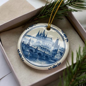 May include: A round, ceramic Christmas ornament featuring a blue and white illustration of a European city scene. The ornament has a gold string for hanging and is presented in a gift box with a sprig of evergreen.