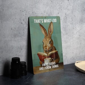 May include: A metal sign with a blue background and a brown rabbit reading a book. The sign says "That's what I do I read books and I know things".