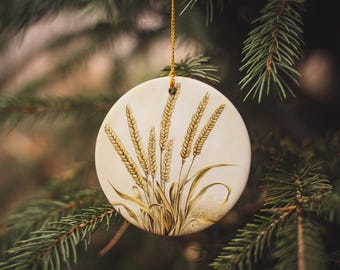 May include: A white ceramic ornament with a hand-painted design of wheat stalks in shades of brown and gold. The ornament is hanging from a gold string.