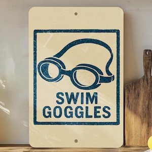 May include: A rectangular sign with a cream-colored background features a blue graphic of swim goggles inside a blue rectangular border. The words "SWIM GOGGLES" are printed in blue below the graphic. The sign is mounted on a wooden surface.