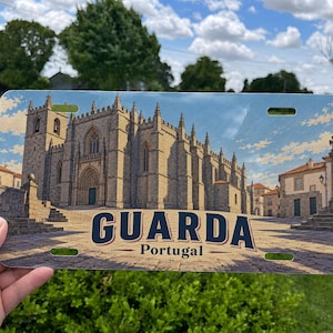 May include: A rectangular metal license plate featuring a printed image of the Cathedral of Guarda in Portugal. The design displays the stone church architecture, a cobblestone plaza, and the text GUARDA Portugal in bold dark blue lettering.