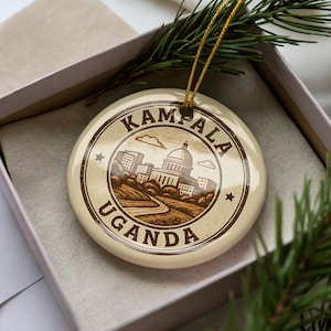 May include: Cream-colored ceramic ornament featuring a detailed illustration of Kampala, Uganda. The design includes a building with a dome, city buildings, and the text "Kampala Uganda" in a circular border. The ornament is in a gift box with a gold string for hanging.