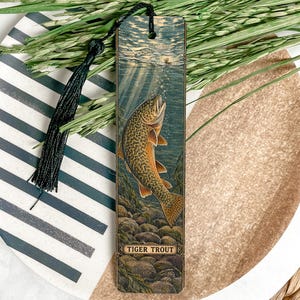 May include: A rectangular wooden bookmark featuring a detailed illustration of a tiger trout swimming underwater among rocks. The bookmark includes a black tassel attached to the top and the text TIGER TROUT printed at the bottom of the design.