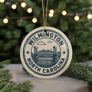 May include: Cream-colored round Christmas ornament with a blue illustration of a bridge and the text "WILMINGTON NORTH CAROLINA". The ornament is hanging from a gold string, with a blurred background of Christmas lights and greenery.
