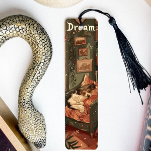 May include: A bookmark featuring a dog sleeping in a chair with a green and brown patterned background. The bookmark has a black tassel and the word "Dream" printed on it.