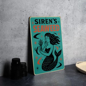 May include: A teal metal sign with the words "SIREN'S SEAWEED" in orange and black. The sign features a black mermaid holding seaweed, with orange crescent moons and stars. The sign is rectangular with rounded corners.
